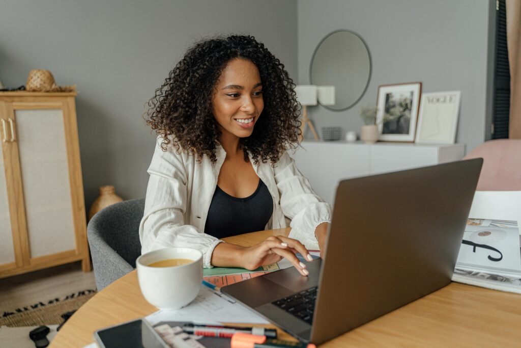 Black woman smiling while working on her laptop at a table, representing a working parent preparing to pump at work.